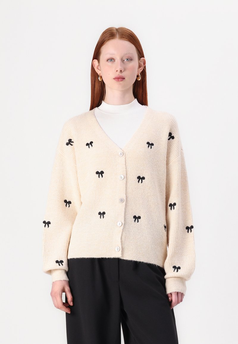 Cardigan de couleur crème avec une texture côtelée, présentant des motifs de nœuds noirs, un col en V et une fermeture à boutons. Porté par-dessus un col roulé blanc.