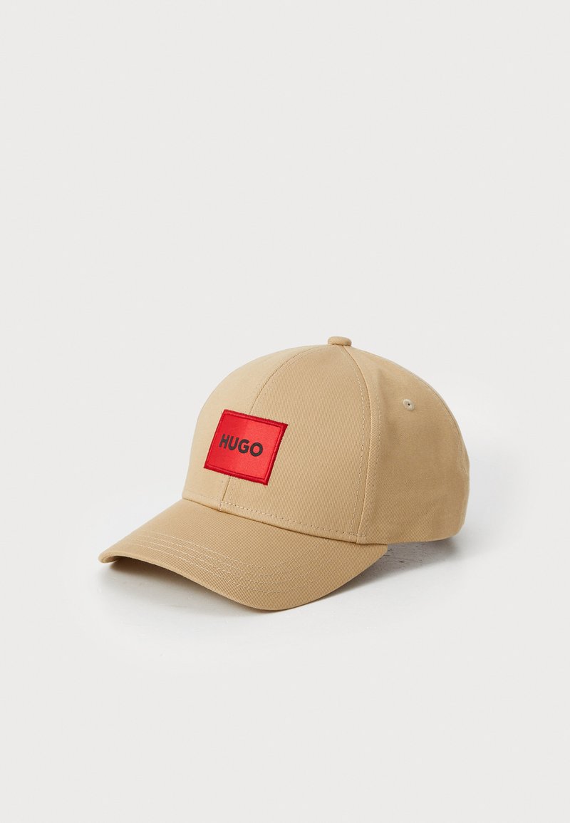 Casquette de base-ball beige en tissu texturé, avec un patch rectangulaire rouge portant "HUGO" en texte noir centré à l'avant.