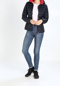 Person som bär en svart puffjacka med röd foder, ljusrosa skjorta, blå tighta jeans och svarta sneakers, stående mot vit bakgrund.