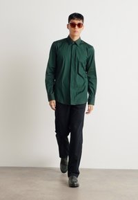 Camisa de manga larga verde, diseño de botones, combinada con pantalones negros y zapatos grises. Gafas de sol transparentes como accesorio. Fondo liso.