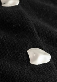 Stříbrný doplněk v organickém tvaru na texturované černé látce, odrážející světlo; na pozadí jsou viditelné černé vlákna.