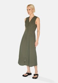 Olivengrünes ärmelloses Kleid mit V-Ausschnitt, vorderem Reißverschluss, Faltendesign und A-Linien-Form, kombiniert mit schwarzen flachen Sandalen.
