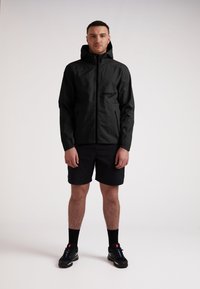 Mand iført en sort hættejakke, sorte shorts, sorte sokker og sorte sneakers, som står imod en ensfarvet hvid baggrund.