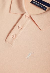 Polo shirt rose clair en tissu tricoté avec un col, trois boutons et un logo brodé blanc sur la poitrine.