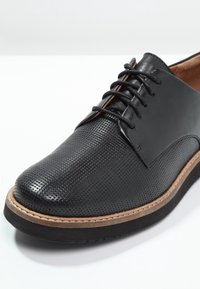 Chaussure en cuir noir avec texture perforée, design à lacets, accent marron sur la semelle et bout lisse et arrondi.