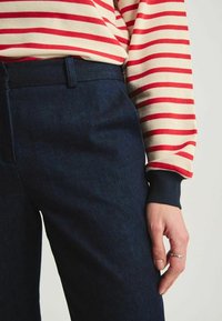 Pantalons en denim noir avec une coupe ajustée et une texture subtile, associés à un haut à manches longues crème et rouge rayé avec des poignets sombres.