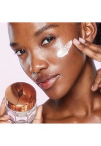 Moisturizer jar with a clear glass base and a reflective rose gold lid. Cream applied to the model's cheek, showcasing a smooth texture.