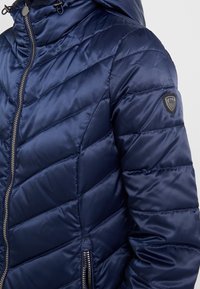 Marinblå puffjacka med en glänsande yta, sicksackmönster, dragkedja framtill och huva. Har en logotyppatch på ärmen.