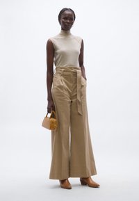 Top bej fără mâneci, pantaloni bej cu talie înaltă, cu croială largă și curea, ținând o geantă mică galbenă, încălțată cu pantofi maro cu toc și vârf ascuțit.