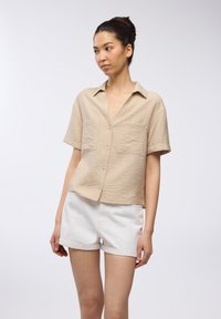 Camicia khaki a maniche corte con chiusura a bottoni e due tasche sul petto. Presenta un tessuto testurizzato e una vestibilità rilassata. Abbinata a pantaloni corti bianchi.