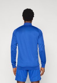 Μπλε αθλητικό τοπ από λείο ύφασμα, με ψηλό γιακά, μακριά μανίκια και λευκές ρίγες στα πλαϊνά. Συνδυάζεται με μπλε σορτς που ταιριάζουν.