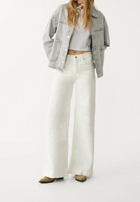 Žena v světle šedé džínové bundě, bílé zkrácené topu, širokých bílých kalhotách s vysokým pasem a hnědých botách s špičatou špičkou, stojí s rukama v kapsách.