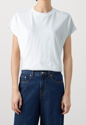 Personne portant un t-shirt blanc uni à manches courtes, rentré dans un jean en denim bleu foncé taille haute avec des rivets en cuivre.