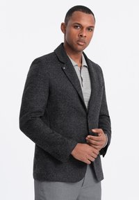 Blazer ajusté gris foncé avec revers et accents de boutons, associé à un polo texturé gris clair en dessous.