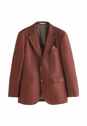 Mann trägt rostfarbenen Anzug und beige Hemd mit Kragen, steht vor neutralem strukturiertem Hintergrund und schaut leicht zur Seite.