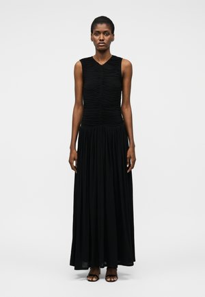 Kobieta stoi na białym tle, ubrana w czarny, bezrękawnik z marszczeniami oraz długą, luźną czarną spódnicę i czarne sandały z odkrytymi palcami.