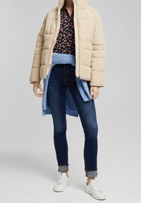 Beige kordfløyels pufferjacka med dragkedja framtill, kombinerad med en blommig blus, blå stickad sjal, mörka denimjeans och vita sneakers.