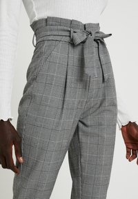 Hoge taille, geruite broek in monochroom grijs materiaal, met een strikdetail op de taille en plooien voor extra textuur.