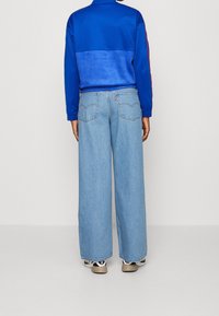 Chaqueta deportiva azul con un diseño de bloques de color, combinada con unos vaqueros de pernera ancha en un azul claro. Logo de Levi’s visible en el bolsillo, llevada con zapatillas.