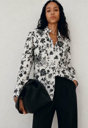 Femme aux longs cheveux bouclés porte un chemisier blanc à fleurs avec des boutons et un pantalon noir, tenant un grand sac noir contre un mur uni.