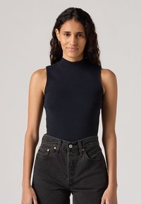 Body nera senza maniche con alto colletto a imbuto, realizzata in tessuto morbido, abbinata a jeans in denim neri a vita alta con accenti in rame.