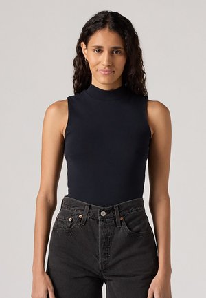 Zwarte mouwloze jumpsuit met een hoge kraag, gemaakt van soepele stof, gecombineerd met hoge zwarte denim jeans met koperen accenten.