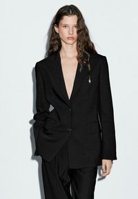 Blazer nero con scollo a V profondo, spalle strutturate e chiusura a un bottone. Presenta una piccola spilla decorativa e una consistenza del tessuto liscia.