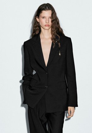 Blazer nero con scollo a V profondo, spalle strutturate e chiusura a un bottone. Presenta una piccola spilla decorativa e una consistenza del tessuto liscia.
