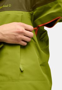 Chaqueta verde de exterior con una sección superior marrón. Presenta una cremallera naranja y una tela texturada. Se ve una mano ajustando la cremallera.