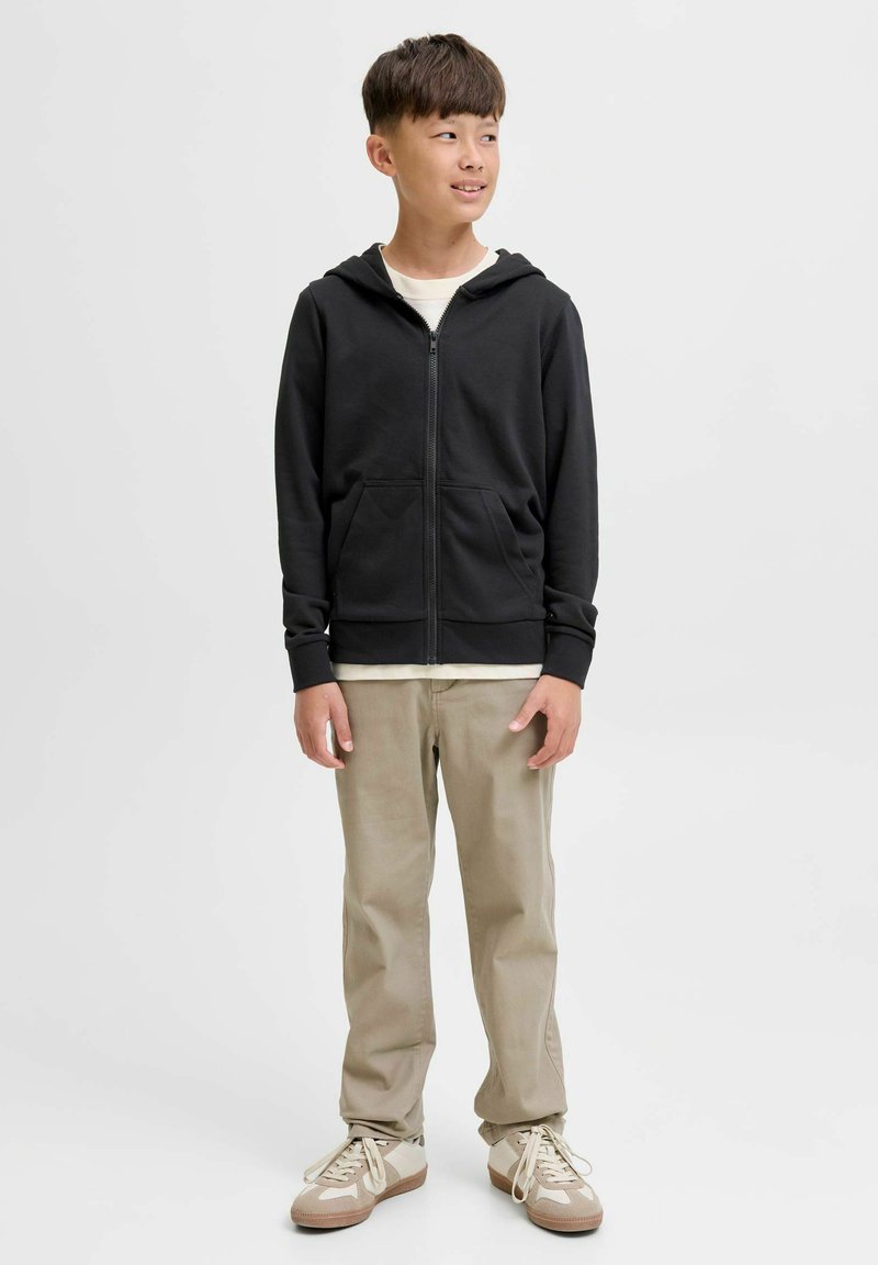 Haut à capuche zippé noir avec des poches kangourou, porté sur une chemise blanche. Un pantalon beige et des sneakers marron complètent la tenue. Texture du tissu lisse.