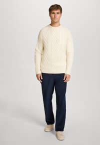 Beiger, strukturiert gestrickter Pullover mit Rundhalsausschnitt und gerippten Bündchen, kombiniert mit dunkelblauen Hosen und hellen Sneakers. Einfaches, texturiertes Design.