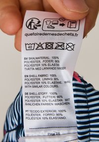 Metka z instrukcjami pielęgnacji w ręku, zawierająca instrukcje pielęgnacji w wielu językach, informująca o materiałach: 100% poliester na zewnątrz, 90% poliester podszewka, 10% elastan.