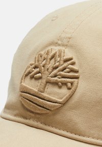Beige baseball cap lavet af stof, med et præget trælogo. Synlige stikninger langs skyggen og toppen.