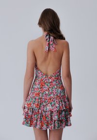 Kukallinen halterneck-mekko puuvillasta, jossa on matala selkäosa, kerroksellisia röyhelöitä ja vaalealla taustalla pinkki-, punainen- ja vihreäkukkojen kuvio.