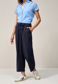 Žena v krátkém rukávu světle modré košili na knoflíky, tmavomodrých širokých kalhotách s páskem na stažení a bílých teniskách s černým logem "V".