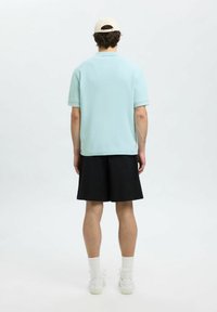 Ljusblå bomullspolo med korta ärmar, parat med svarta shorts och vita sneakers, sedd från baksidan mot en vit bakgrund.