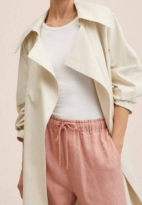 Crème trenchcoat met een brede kraag over een witte geribde tanktop; gecombineerd met lichtroze elastische broek met taille. Zachte, relaxte pasvorm.