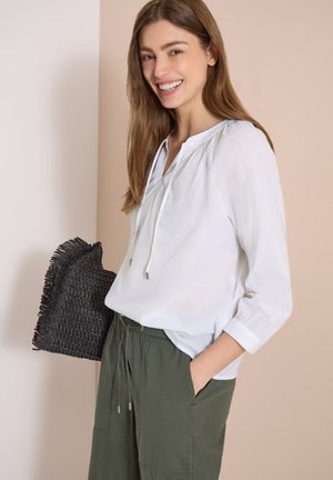 Lächelnde Frau in weißer Bluse und grüner Hose hält eine schwarze, strukturierte Clutch vor neutralem Hintergrund.
