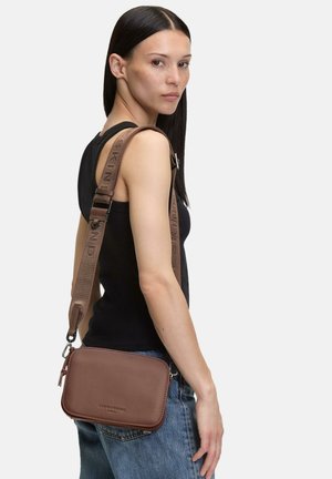 Frau mit glatten dunklen Haaren trägt ein schwarzes ärmelloses Oberteil und Jeans, sie trägt eine braune Crossbody-Tasche mit einem breiten Riemen über der Schulter.