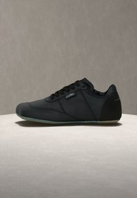 Μαύρο χαμηλό σνίκερ με κορδόνια, λείο επάνω μέρος και ελαφρώς διαφανές πέλμα, εμφανίζεται σε ουδέτερο φόντο πάνω σε επίπεδη επιφάνεια.