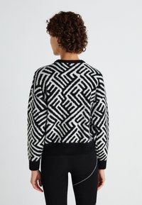 Pull en tricot noir et blanc avec un motif géométrique, col rond et poignets et ourlet côtelés ; texture douce. Porté avec des leggings noirs.