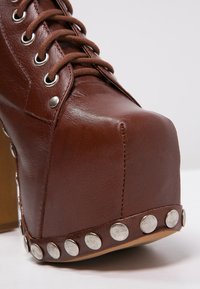 Zapato de plataforma de cuero marrón con punta cuadrada, tacón elevado y tachuelas circulares plateadas a lo largo de la base. Presenta ojales con cordones y superficie texturizada.