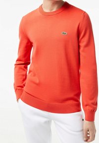 Czerwony sweter z okrągłym dekoltem, ribbowanymi mankietami i dołem. Posiada małe zielone logo krokodyla na klatce piersiowej. Miękka dzianinowa tekstura.