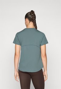 Mulher com cabelo longo trançado, vestindo uma camisa de mangas curtas teal e leggings castanhas, vista de costas contra um fundo simples.