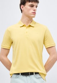 Jeune homme portant un polo à manches courtes jaune clair et un jean bleu clair, debout les mains dans le dos, regardant sur le côté.