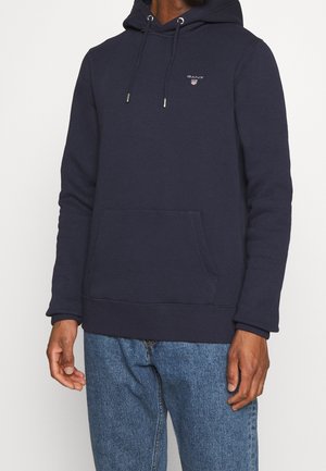 Man wearing navy blue GANT hoodie with front pocket and light blue jeans against plain white background.