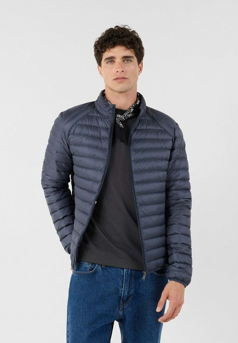 Giacca imbottita blu navy con compartimenti orizzontali, chiusura con zip e colletto alto. Indossata sopra una maglia grigio scuro e jeans, presenta una lucentezza sottile.