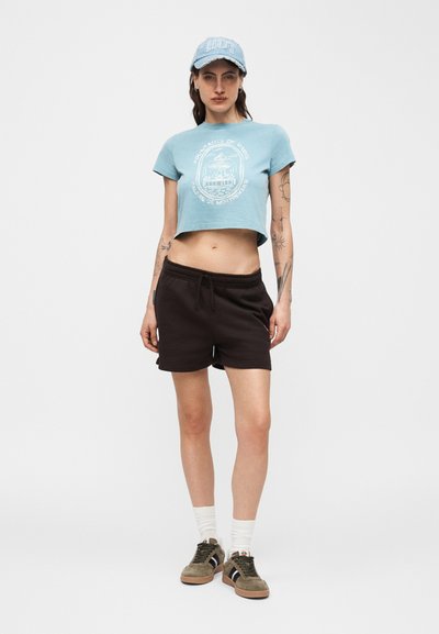 Νεαρό άτομο όρθιο, φοράει ανοιχτό μπλε κοντομάνικο μπλουζάκι με στάμπα, μαύρα σορτς, ανοιχτό μπλε καπέλο, λευκές κάλτσες και αθλητικά παπούτσια, μπροστά σε απλό φόντο.