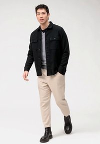 Giacca nera con bottoni sopra un maglione grigio, pantaloni beige a coste e stivaletti neri alla caviglia. L'outfit presenta una vestibilità rilassata e colori neutri.