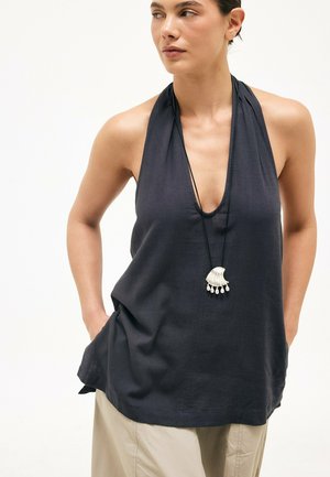 Femme portant un haut dos nu sombre avec un décolleté en V profond et un collier pendentif, mains dans les poches, regardant vers sa gauche.
